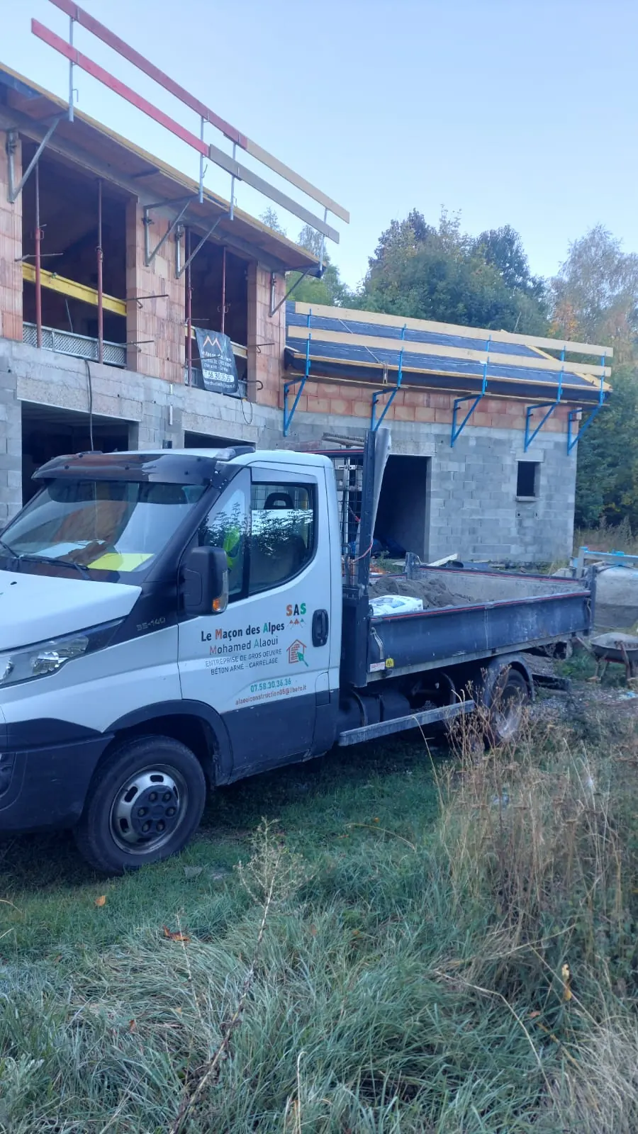 Camion-benne floqué Le Maçon des Alpes garé devant une maison en construction, rez-de-chaussée en parpaings et étage en brique
