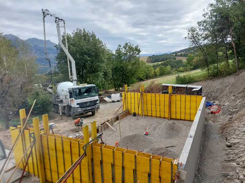 Camion-pompe à béton devant des banches de coffrage jaunes, sur un chantier en pente face aux montagnes