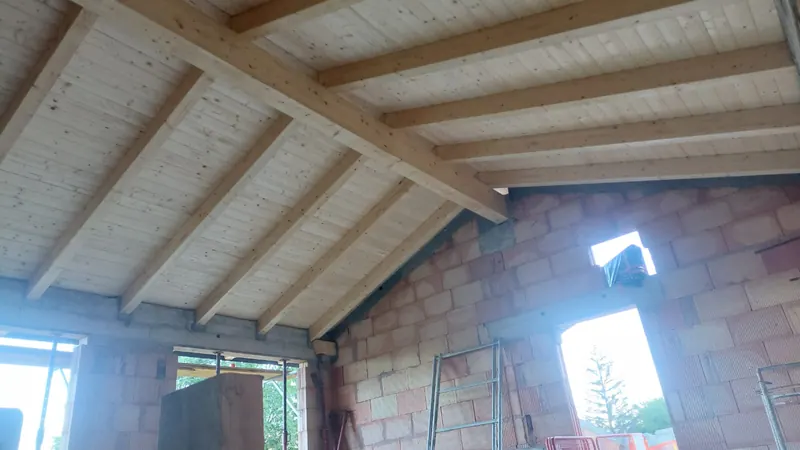 Intérieur d'une maison en construction : murs en brique et chaînages béton sous une charpente en bois